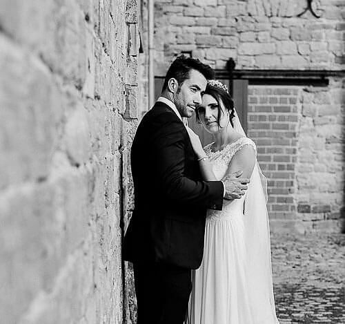 Paar lehnt an einer Mauer. Sie sind sich zugewendet und Braut schaut in Richung Fotograf