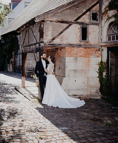 Bräutigam lehnt an einem Pfosten eines alten Fachwerkhauses und hat seine Braut im Arm